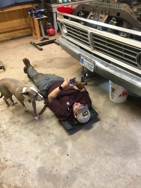 1994 Ford Bronco - A man lies on a creeper under the front of a classic truck, with a dog nearby.