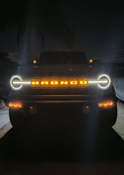 2023 Ford Bronco - The front grille of a 2023 Ford Bronco features illuminated lettering and distinctive halo headlights, visible at night.