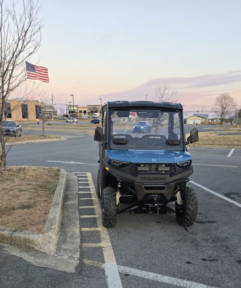 2025 Polaris RANGER - A blue Polaris UTV is parked in a lot with an American flag flying nearby.