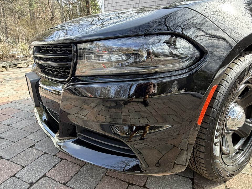Front corner of black Dodge Charger with grille and headlight visible. Tire has 'ASSURANCE' text