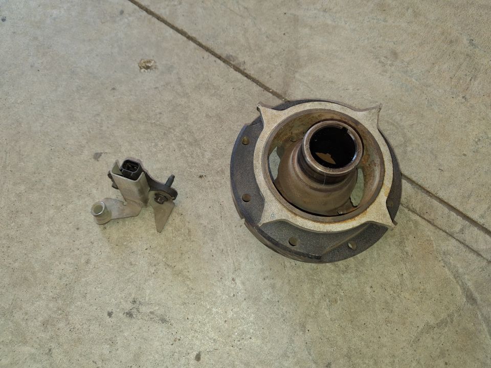 Two automotive parts on a concrete floor: a metal sensor with a black connector and a circular metal component with a central opening