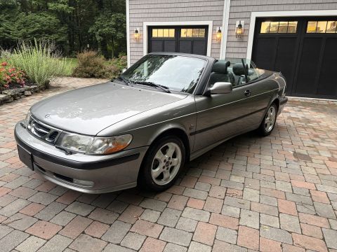 2001 Ford Crown Victoria - Front three-quarter view of a silver-gray Saab 9-3 convertible with the top down, parked on a brick paver driveway in front of a shingled h…