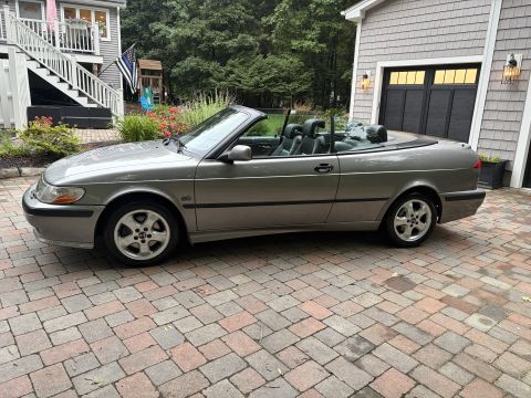2001 Ford Crown Victoria - Side profile of a pewter-gray Saab convertible with the top down, parked on a brick paver driveway in front of a gray shingled house and ga…