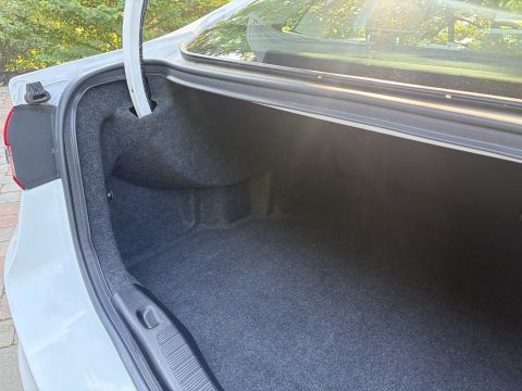 2001 Ford Crown Victoria - Open, empty trunk of a white sedan lined with dark gray carpet, showing the trunk hood, weatherstrip and a red taillight edge. Green foliag…