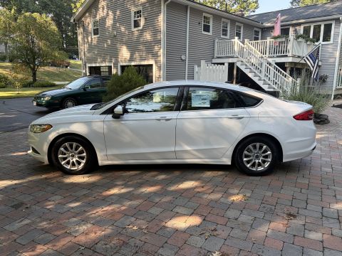 2001 Ford Crown Victoria - Side profile of a white Ford Fusion sedan parked on a brick paver driveway, with a dealer window sticker on the rear glass; a green sedan a…
