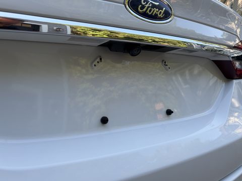 2001 Ford Crown Victoria - Close-up of the rear deck lid of a white Ford, showing the chrome Ford oval badge, a trunk-mounted backup camera in the chrome trim strip,…