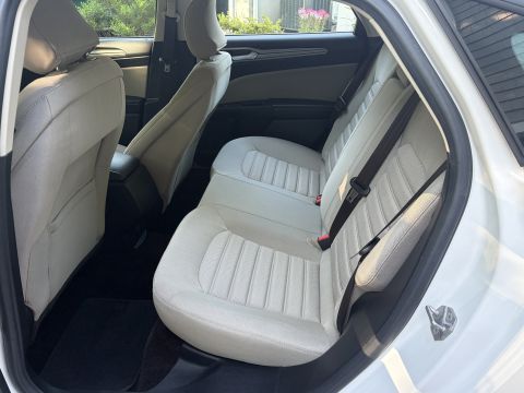 2001 Ford Crown Victoria - Rear bench seat of a white sedan viewed through the open rear door, upholstered in light tan/gray cloth with black seatbelts and dark carpe…