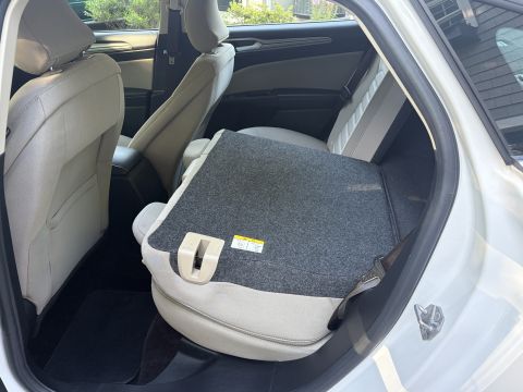 2001 Ford Crown Victoria - Rear seat of a white sedan with the center armrest folded down, upholstered in light tan cloth; the open rear door and dark floor mats are…