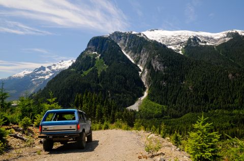 1985 Ford Bronco - A lifted blue Ford Bronco with the top removed sits at the edge of a gravel logging road, seen from behind, overlooking a forested valley a…