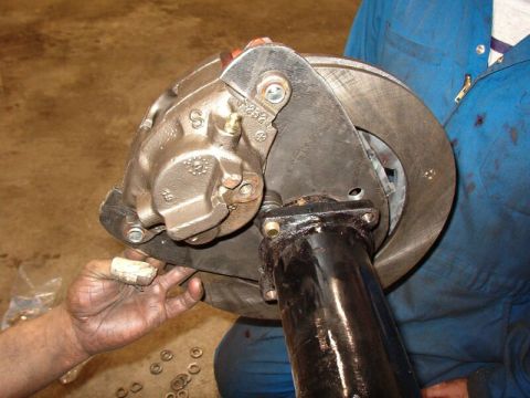 1985 Ford Bronco - A close-up view shows a person's hands working on a brake caliper and rotor assembly attached to a black metal axle tube. The brake caliper…