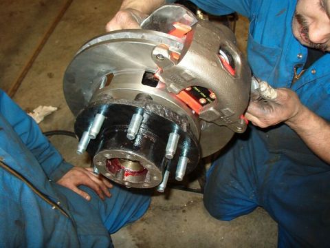 1985 Ford Bronco - A mechanic in blue overalls is working on a brake caliper and rotor assembly, with red brake pads visible inside the caliper. The rotor is…
