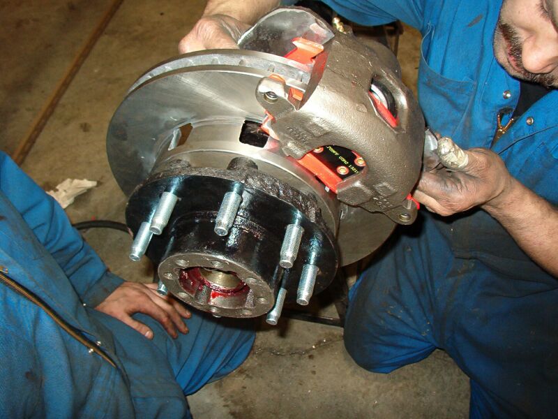 Close-up of a vehicle brake caliper and rotor assembly with red brake pads and greased lug studs.