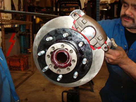 1985 Ford Bronco - A mechanic is installing a brake caliper over a rotor and hub assembly. The hub is greased and has several lug nuts installed.