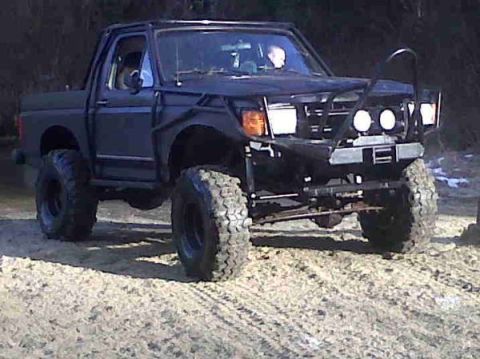 1985 Ford Bronco - A dark colored, modified Ford Bronco is shown from a three-quarter front angle, parked on a dirt or sand surface. The vehicle features larg…