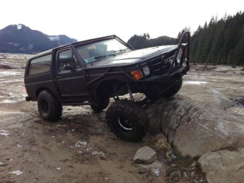 1985 Ford Bronco - A modified black Ford Bronco is navigating over large rocks in a muddy, off-road environment with mountains and trees in the background.