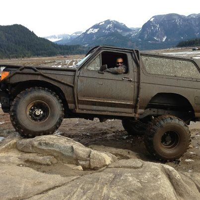 1985 Ford Bronco - A heavily modified, mud-covered 1980s Ford Bronco is shown navigating a rocky terrain with a driver behind the wheel. The vehicle features…