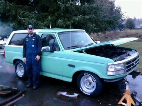 1985 Ford Bronco - A man in blue overalls stands next to a light green Ford Bronco with its hood open. The vehicle is parked on a wet surface with trees in th…