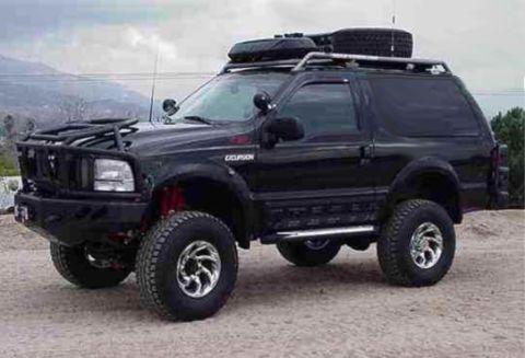 1985 Ford Bronco - A black, lifted Ford Excursion SUV is parked on a dirt lot with a mountainous landscape in the background. The vehicle features a custom fr…