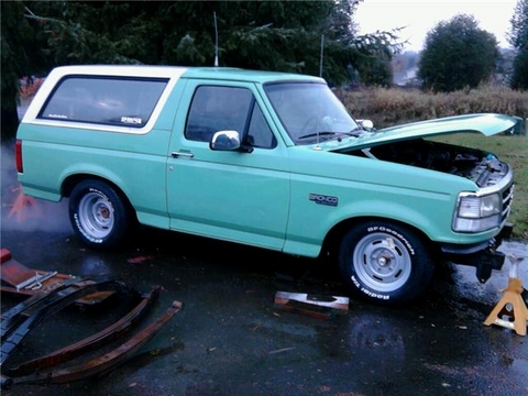 1985 Ford Bronco - A teal and white Ford Bronco is parked on a wet surface with its hood open. Several leaf springs are laid out on the ground in front of the…