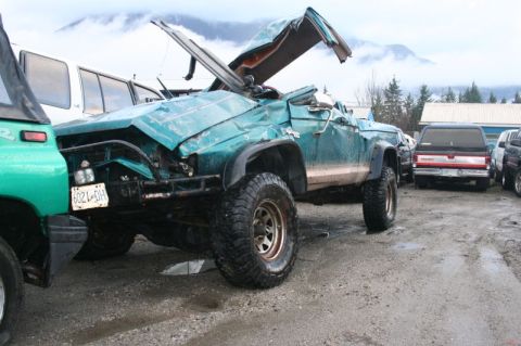 1985 Ford Bronco - A severely damaged teal Jeep Cherokee is parked in a lot with other vehicles, mountains in the background. The roof and hood are heavily cr…
