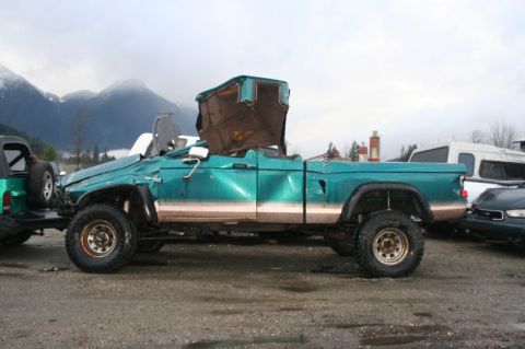 1985 Ford Bronco - A heavily damaged teal pickup truck with its roof section ripped open and bent upwards is parked in a gravel lot with mountains in the back…