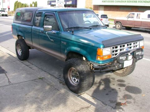 1985 Ford Bronco - A lifted, teal Ford Ranger pickup truck with a camper shell is parked on a street next to a sidewalk. The truck features large off-road tir…