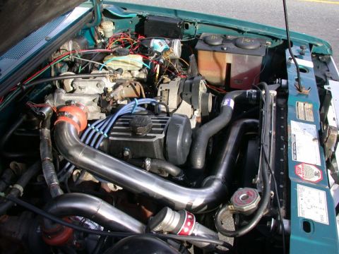1985 Ford Bronco - The engine bay of a green Ford Bronco is shown, featuring a modified intake system with large polished pipes and red silicone couplers. Var…