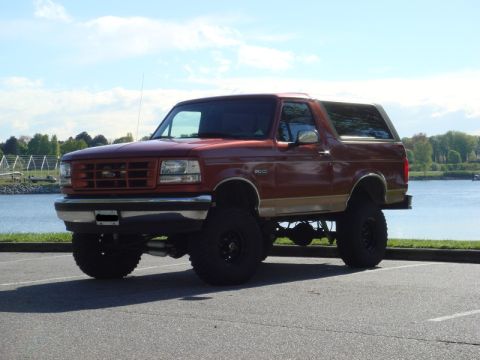 1985 Ford Bronco - A lifted, dark red 1980s Ford Bronco is parked on asphalt next to a body of water. The vehicle has large off-road tires and a tan accent st…