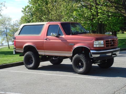 1985 Ford Bronco - A lifted, two-tone red and tan Ford Bronco is parked in a lot with trees and a grassy area in the background. The SUV has large off-road ti…