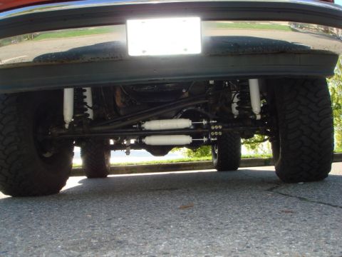 1985 Ford Bronco - The underside of a lifted Ford Bronco is shown, featuring large off-road tires, white shock absorbers, and a visible steering linkage.