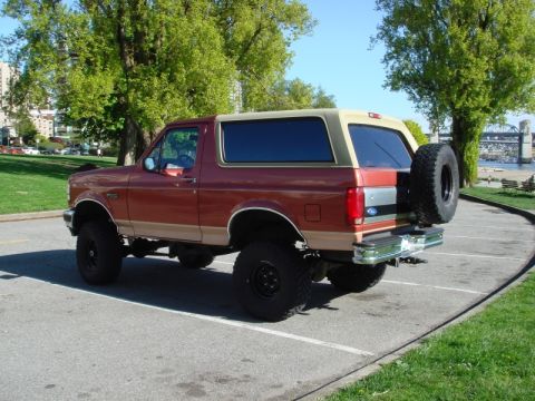 1985 Ford Bronco - A lifted, two-tone red and tan Ford Bronco is parked in a paved lot with a grassy area and trees in the background. The rear-mounted spare…