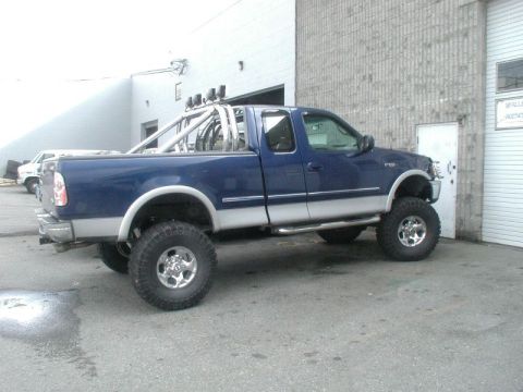 1985 Ford Bronco - A lifted, dark blue and silver pickup truck with a chrome roll bar and auxiliary lights is parked on asphalt in front of a building.
