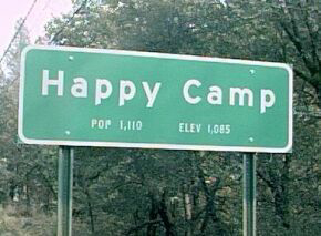 1985 Ford Bronco - A green sign reads "Happy Camp" with "POP 1,110" and "ELEV 1,085" below it. The sign is mounted on two poles against a backdrop of trees.