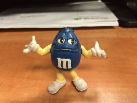 1985 Ford Bronco - A blue M&M candy figurine with white shoes and yellow arms is standing on a wooden surface. The figurine has the letter "m" on its chest an…
