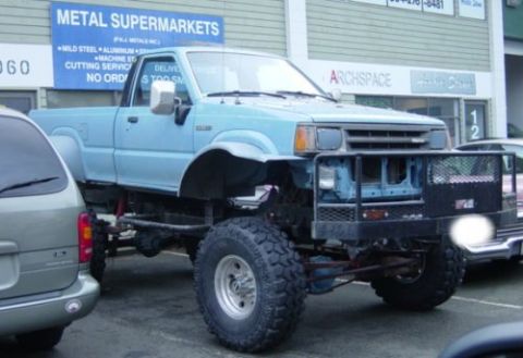 1985 Ford Bronco - A lifted, light blue pickup truck with large off-road tires is parked in front of a building with a "METAL SUPERMARKETS" sign. The front of…