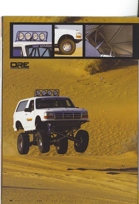 1985 Ford Bronco - A lifted white Ford Bronco is captured mid-air, jumping over sand dunes. The vehicle features a custom roll cage, large off-road tires, and…
