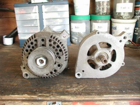 1985 Ford Bronco - Two automotive alternators are displayed on a wooden workbench. The alternator on the left is a complete unit with a pulley, while the one…
