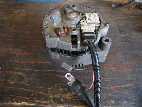 1985 Ford Bronco - A close-up, overhead view of a vehicle alternator with wiring harness connected and a battery cable lying nearby on a wooden surface.