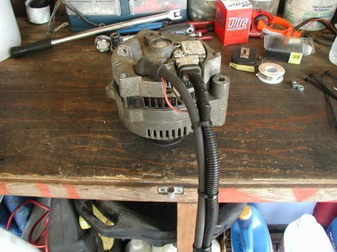 1985 Ford Bronco - A car alternator is shown on a wooden workbench with various tools and supplies, including a roll of solder, zip ties, and a red box labele…