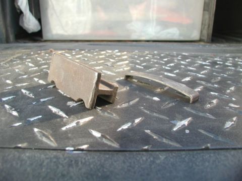 1985 Ford Bronco - Two metal parts are lying on a black diamond plate surface. One part is a T-shaped metal piece, and the other is a curved metal strip.