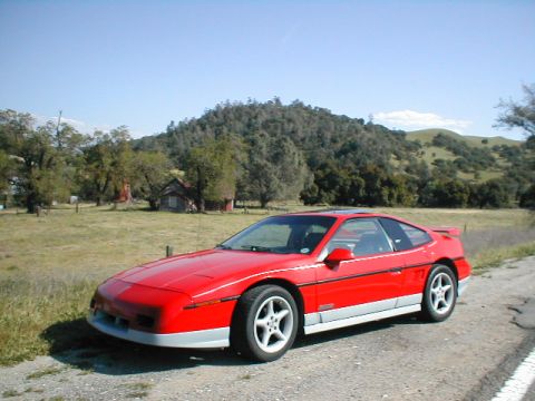 1986 Pontiac Fiero - A red Pontiac Fiero is parked on a dirt road, with a house and hills in the background.