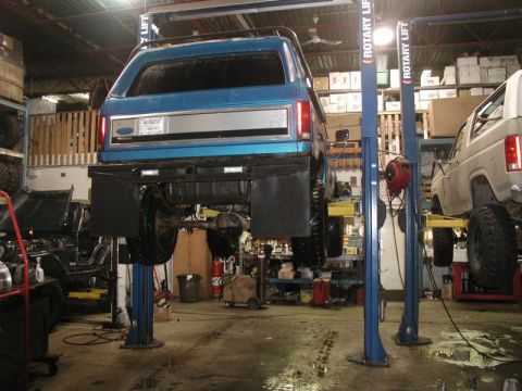 1985 Ford Bronco - The rear of a blue Ford Bronco is shown lifted on a two-post automotive lift in a garage. Another white SUV is visible on a lift to the rig…