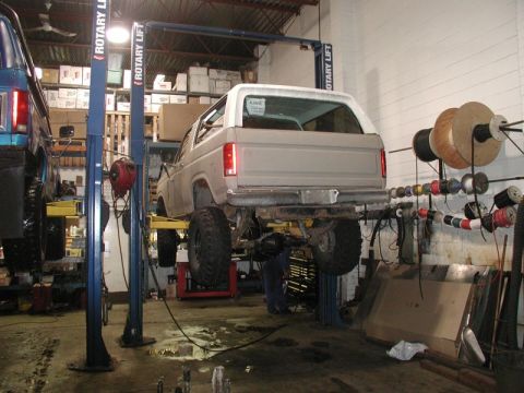 1985 Ford Bronco - A light-colored Ford Bronco is raised on a two-post rotary lift in a workshop. The rear of the vehicle is visible, showing large off-road t…