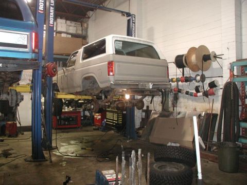 1985 Ford Bronco - A light-colored SUV is raised on a hydraulic lift in a garage, with its underside exposed for maintenance. Another blue SUV is visible on a…