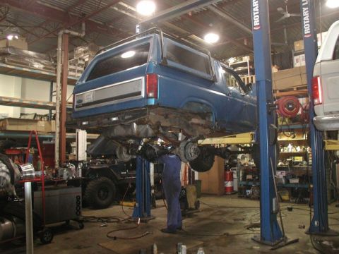 1985 Ford Bronco - A blue Ford Bronco is raised on a hydraulic lift in a garage with a mechanic working underneath it. The vehicle is viewed from the rear qua…