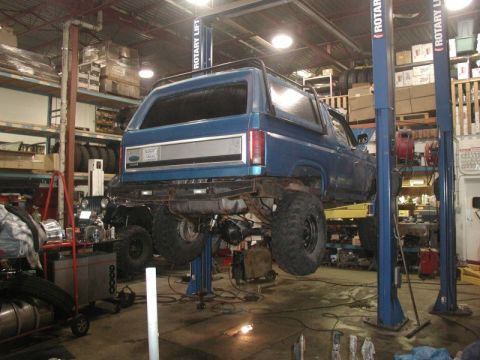 1985 Ford Bronco - A blue Ford Bronco is raised on a two-post car lift in a garage. The rear of the vehicle is visible, showing large off-road tires and a mod…