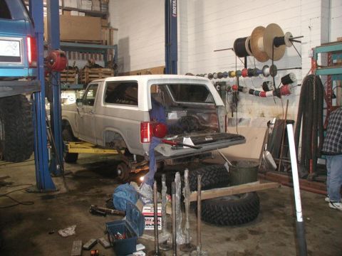 1985 Ford Bronco - A gray Ford Bronco is on a lift in a garage with its rear tailgate open. Various tools and parts are scattered on the floor around the vehi…