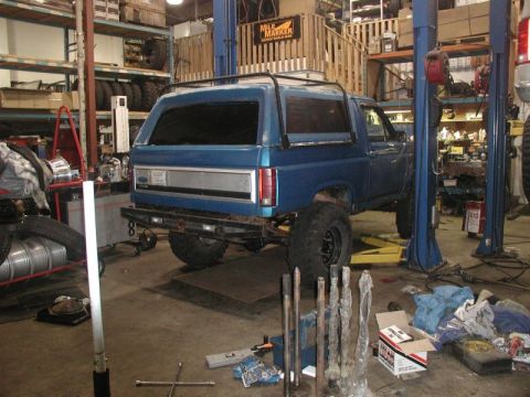 1985 Ford Bronco - A blue Ford Bronco is parked in a garage with its rear end raised on a hydraulic lift. Several metal shafts are laid out on the floor in fr…
