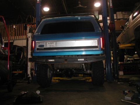 1985 Ford Bronco - The rear of a blue Ford Bronco is shown lifted on a car lift in a garage. The vehicle has a custom rear bumper and large off-road tires.