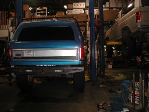 1985 Ford Bronco - The rear of a blue Ford Bronco is shown in a garage, lifted on a car lift. Another white Ford Bronco is visible on a separate lift in the b…